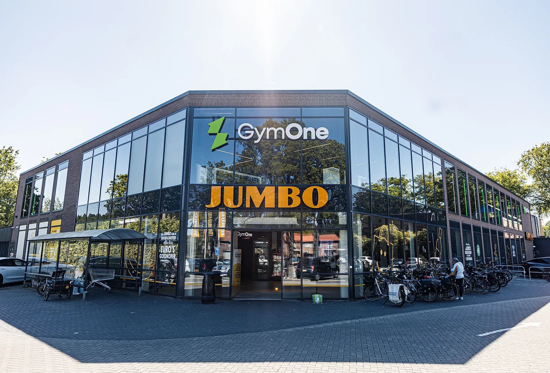 Exterieur van GymOne Emmen: hoekgebouw met GymOne-logo en glazen gevel boven de JUMBO