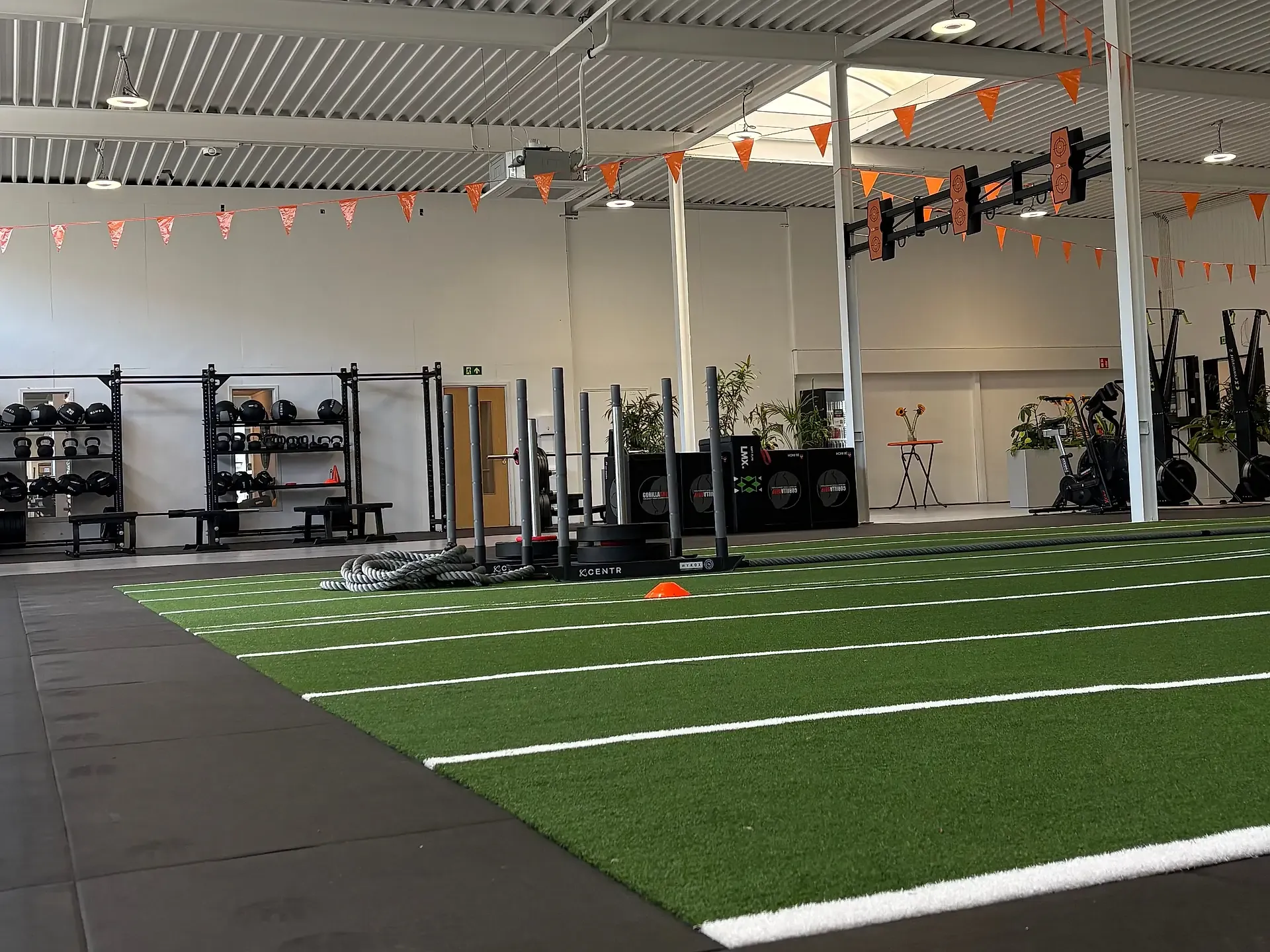 Lifestyleclub360 Drachten: HYROX-ruimte met kunstgras-sprintbaan, sleds, wall-ball-rig en cardiozone onder oranje vlaggenlijnen